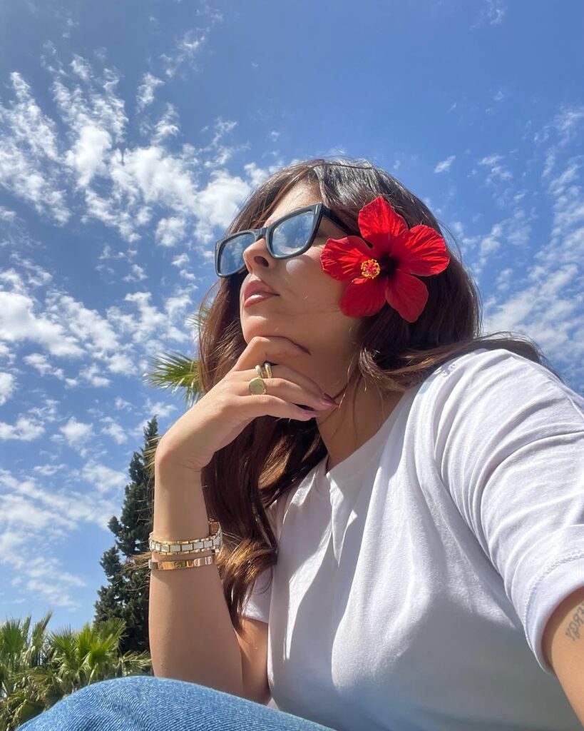 Woman wearing sunglasses and a white shirt with a red hibiscus flower behind her ear, sitting under a bright blue sky with scattered clouds and palm trees in the background.