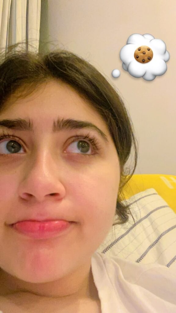 Girl lying in bed with a funny pout, looking up toward a cookie emoji in a thought bubble above her head.