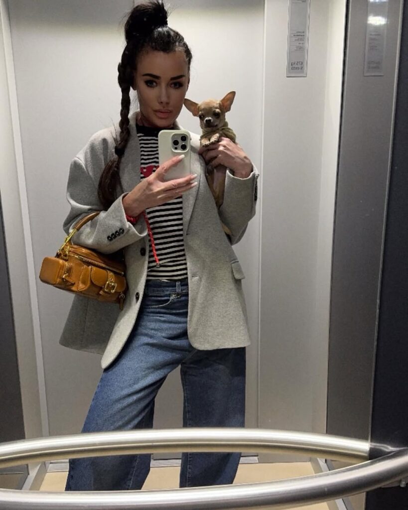 Woman taking a mirror selfie in an elevator, holding a small chihuahua, wearing a grey blazer over a striped shirt with wide-leg jeans and a mustard handbag.